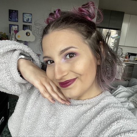 Girl with tan skin, purple hair and makeup, and a light grey cropped sweater.